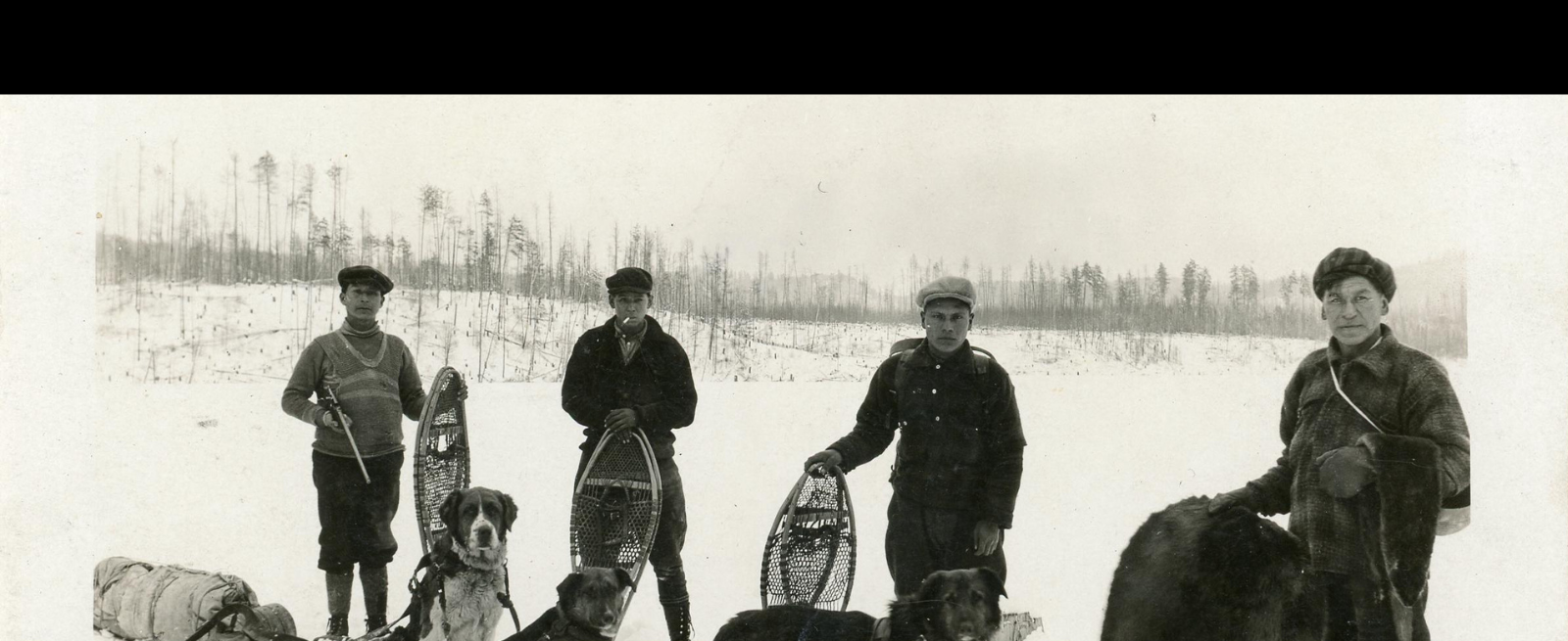 Four fur traders standing in snow covered landscape with snowshoes and team of dogs